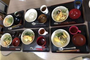 介護施設の食事風景。複数人分の栄養バランスの取れた食事が黒いトレーに配膳されている。